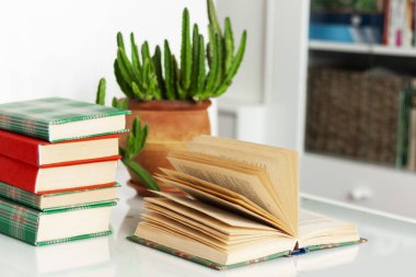 Konforlu ev dekorasyonu: kitap yığını, saksıda bitkiler, beyaz bir masanın üzerinde açık bir kitap. Uzak ev eğitimi. Evde kalma karantina kavramı.