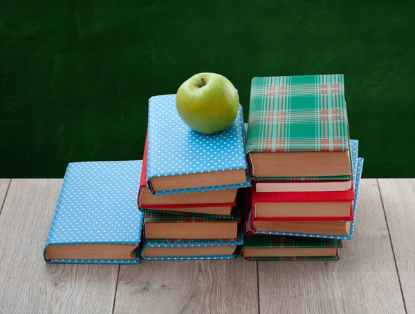 Back to school, pile of books in colorful covers and green apple on wooden table with empty green school board background. Distance home education. Quarantine concept of stay home