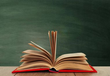 Ahşap bir masanın üzerinde, yeşil bir okulun kara tahtasının üzerinde kırmızı bir kapakta açık bir kitap kitabı. Okul mesafesindeki ev eğitimine geri dönüyoruz. Evde kalma karantina kavramı.