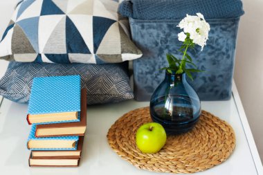Konforlu ev dekorasyonu: bir yığın kitap, yeşil elma, dekoratif yastıklar, ekoseli mavi kutu ve cam bir masada beyaz çiçekli vazo. Uzak ev eğitimi. Evde kalma karantina kavramı.