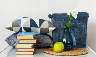 Konforlu ev dekorasyonu: bir yığın kitap, yeşil elma, dekoratif yastıklar, ekoseli mavi kutu ve cam bir masada beyaz çiçekli vazo. Uzak ev eğitimi. Evde kalma karantina kavramı.