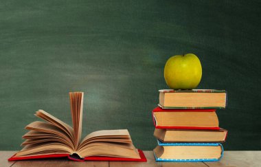 Açık kitap kitabı, bir yığın kitap ve yeşil okul karatahtasının arka planındaki ahşap masanın üzerindeki yeşil elma. Okul mesafesindeki ev eğitimine geri dönüyoruz. Evde kalma karantina kavramı.