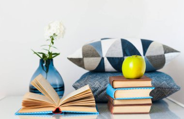 Konforlu ev dekorasyonu: Bir yığın kitap, yeşil elma, dekoratif yastıklar ve cam bir masanın üzerinde beyaz çiçekli vazo. Uzak ev eğitimi. Evde kalma karantina kavramı.