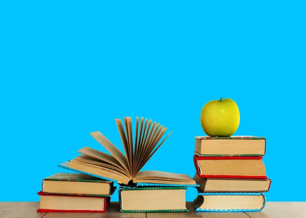 Open textbook, pile of books in colorful covers and green apple on wooden table with blue background. Distance home education. Back to school, quarantine concept of stay home.