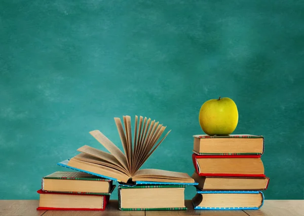 Open textbook, pile of books in colorful covers and green apple on wooden table with green blackboard background. Distance home education. Back to school, quarantine concept of stay home
