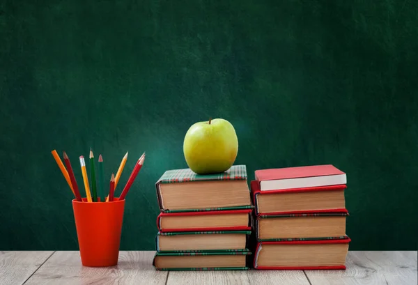 pile of books in colorful covers, pencils in holder and green apple on wooden table with green blackboard background. Distance home education. Back to school, quarantine concept of stay home