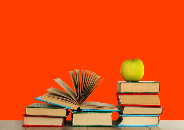Open textbook, pile of books in colorful covers and green apple on wooden table with red orange background. Distance home education. Back to school, quarantine concept of stay home