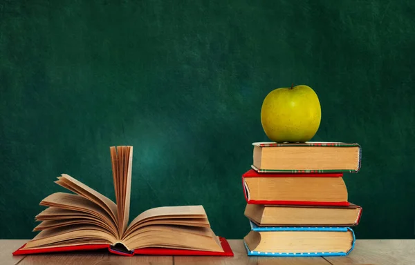 Open book textbook, pile of books and green apple on a wooden desk on the background of a green school chalkboard. Back to school distance home education.Quarantine concept of stay home