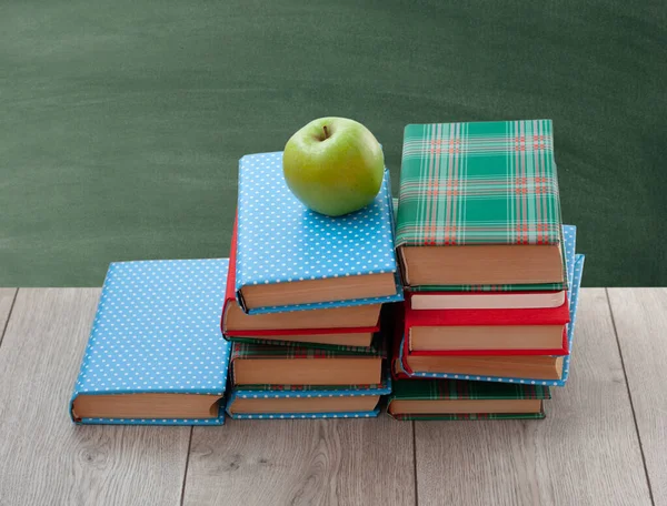 Back to school, pile of books in colorful covers and green apple on wooden table with empty green school board background. Distance home education. Quarantine concept of stay home