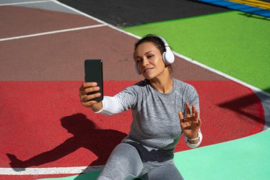 Gri eşofmanlı genç kadın yeşil bir mindere oturmuş kulaklık takıyor ve video iletişimiyle iletişim kuruyor.