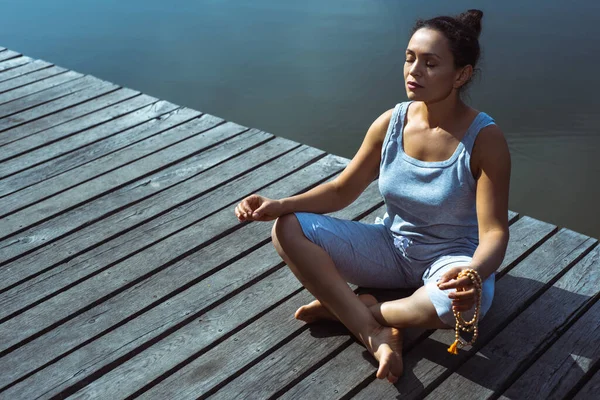 The young woman on a wooden pier sits in a lotus position with her eyes closed. Meditation, yoga in nature.