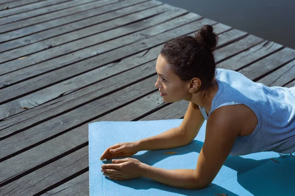 The young woman does a plank exercise on a gymnastic mat and looks in front of her. Side view. Meditation, yoga in nature.