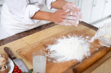 Aşçının ellerine yakın çekim yap. Aşçı ellerindeki unu masaya vurarak eziyor. Masanın üzerinde dağınık un ile ahşap tahta var. Yan görünüm.