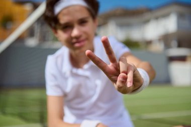 Spor kıyafetleri giyen genç bir tenis meraklısı sahada güneşli bir günün tadını çıkarırken barış işareti gösteriyor. Parlak arka plan ve jestlere odaklanma pozitifliği ve enerjiyi iletir.
