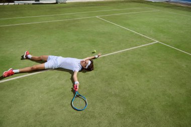Zorlu bir maçtan sonra bitkin düşmüş bir tenisçi çimen kortunda yatıyor. Görüntü, kararlılık ve sevinç duygularını yakalar. Canlı yeşiller ve tenis kıyafetleri sahneyi hazırladı..