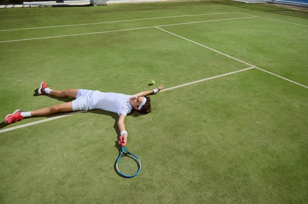 Zorlu bir maçtan sonra bitkin düşmüş bir tenisçi çimen kortunda yatıyor. Görüntü, kararlılık ve sevinç duygularını yakalar. Canlı yeşiller ve tenis kıyafetleri sahneyi hazırladı..