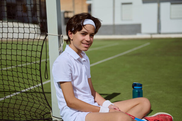 A young tennis athlete rests sitting on a tennis court after a session, dressed in white attire with gear and a drink bottle visible. This depicts the determination and sportsmanship of youth athletes.