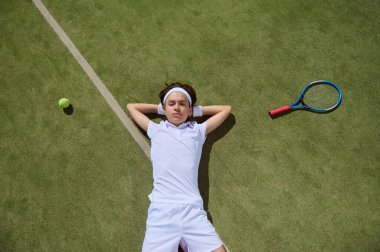 Beyaz elbiseli bir tenisçi, elinde raket ve topla çimenli bir kortta dinleniyor. Spor ortamında huzurlu ve huzurlu bir atmosfer, rahatlama, sportmenlik ve rahatlık kavramları için mükemmel..