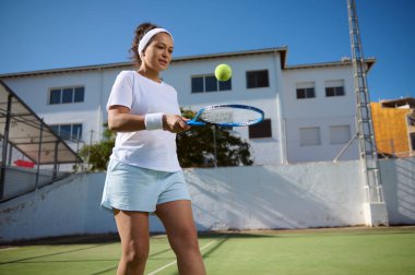 Tenis kıyafeti giymiş bir kadın raketinde top sektiriyor. Açık hava tenis kortunda güneşli bir arka planda yakalanan bu sahnede aktif spor katılımı ve boş zamanların vurgulanması öngörülüyor..
