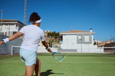 Kendini işine adamış bir bayan tenisçi, açık mavi gökyüzü altında, güneşli bir ortamda, oyuna enerji, yetenek ve tutku sunan, iyi korunan bir açık hava sahasında aktif olarak oyuna katılır..