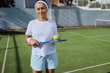 Orta yaşlı bayan bir tenisçi rahat bir kıyafetle açık hava kortunda güvenle duruyor. Sahne, sağlıklı ve aktif yaşama dikkat çeken fitness ve tenis tutkusunu tasvir ediyor..