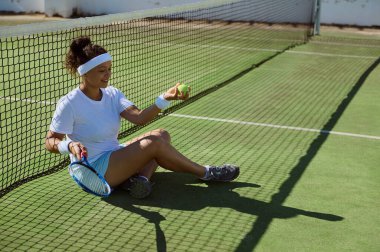 Bir tenisçi, raketini ve topunu tutan bir kortun ağlarının yanında dinleniyor. Rahatlamış ve mutlu görünüyor, güneşli havalarda açık hava sporlarından sonra biraz eğleniyor..