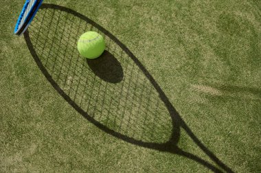 Tenis raketi ve top canlı bir çimen kortuna gölge düşürüyor güneşli bir günde sporun özünü gözler önüne seriyor..