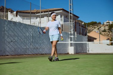 Tenis raketi ve top tutan atletik giyinmiş bir kadın güneşin altında tenis kortunda. Ortam temiz bir gökyüzü ve sakin bir yerleşim yeri içeriyor..