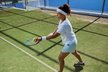 Beyaz elbiseli genç bir kadın çimen kortunda tenis oynuyor, odaklanarak bir çekim hazırlıyor..