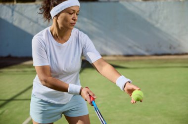 Beyaz elbiseli bir kadın güneşli bir tenis kortunda hizmet etmeye hazırlanıyor. Elinde bir tenis raketi ve top tutuyor, açık ve parlak bir spor ortamında antrenmanına odaklanıyor..