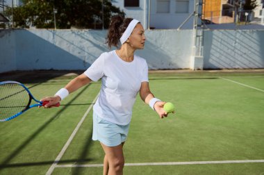 Kendinden emin bir kadın güneşli bir günde tenis antrenmanı sırasında servis yapmaya hazırlanıyor. Spor kıyafetleri içinde, tenis raketi ve top tutarak, odaklandığını ve spora kendini adadığını gösteriyor..