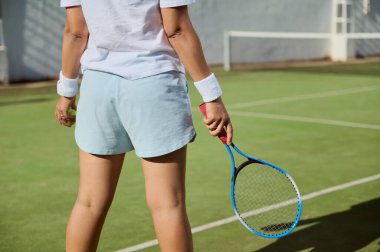 Parlak güneş ışığı altında çimenli bir kortta oynamaya hazır raket tutan bir tenisçinin yakın plan dikiz görüntüsü. Görüntü sporun odak, enerji ve rekabetçi ortamını vurguluyor.