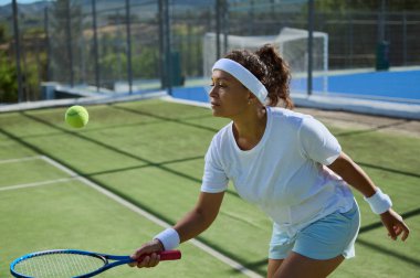 Odaklanmış bayan tenisçi dışarıda sahada topa vurmaya hazırlanıyor. Spor kıyafetleri içinde ve tenis raketi kullanarak çevikliğini ve konsantrasyonunu güneşli bir ortamda gösteriyor..