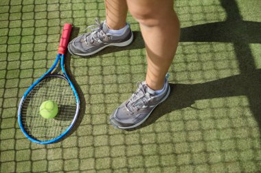 Tenis raketi ve topunun yakın plan görüntüsü bir oyuncunun bacaklarının yanındaki yeşil kortta uzanmış, parlak güneş ışığı ve vurgu için gölgeler eşliğinde bir antrenman veya maç öneriyor.