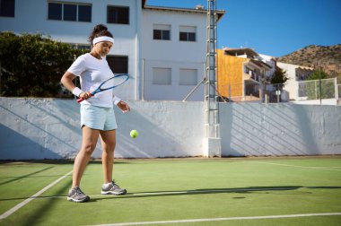 Spor kıyafeti giymiş bir kadın açık hava sahasında tenis oynuyor. Binalarla çevrili, açık havada aktif ve güneşli bir günün tadını çıkarıyor..