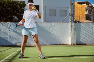 Güneşli bir günde tenis antrenmanı yapmaya hazırlanan odaklanmış bayan sporcu. Bir tenis topu ve raket tutuyor, açık hava sahasında duruyor. Şehir arkaplanı görünür.