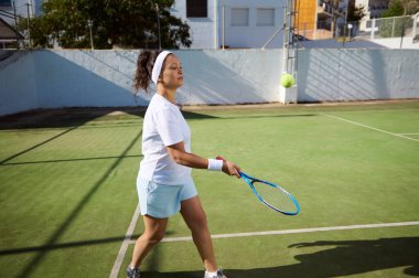 Sportif genç bir kadın güneşli bir günde açık hava kortunda tenis çalışırken konsantre olur. Kararlı ve odaklanmış görünüyor, bir solo çalışma sırasında atletizmini gösteriyor..