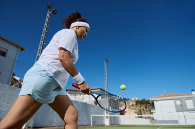 Tenis oynayan bir bayan oyuncu, açık hava sahasında orta sahada yakalandı. Güneş ışığı manzarayı aydınlatıyor, spor kıyafetini ve uzaktaki yerleşim alanını vurguluyor..