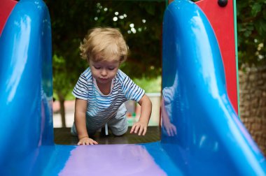 Genç bir çocuk canlı bir oyun parkında renkli bir kaydırağın üzerinde sürünerek tırmanışa odaklanıyor..