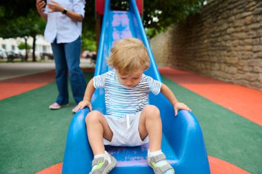 Küçük bir çocuk, bir yetişkin tarafından denetlenirken çocuk parkında kaymaktan zevk alır. Renkli ve canlı ortam açık hava aktiviteleri ve oyun için eğlenceli ve güvenli bir ortam sağlıyor..