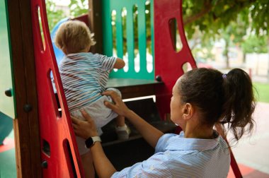 Şefkatli bir anne bebeğine açık havada renkli bir oyun bahçesine tırmanması için yardım eder. Destek, eğlence ve ebeveyn rehberliğini vurgular..