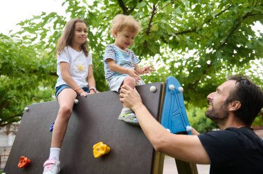 Bir baba, açık havada oyun bahçesine tırmanmayı, güneşli bir günde aile bağlarını ve fiziksel aktivitelerini desteklerken çocuklarını cesaretlendirir ve geçindirir..