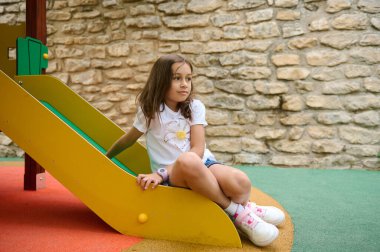 Genç bir kız renkli bir oyun parkı kaydırağında oturuyor. Rahat ve neşeli bir ortamda, taş bir duvarın arkasında düşünceli görünüyor..