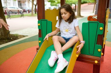 Gülümseyen genç bir kız ağaçlarla çevrili neşeli bir oyun parkında, parlak sarı bir slaytta süzülüyor..