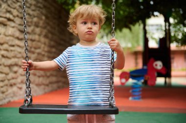 Genç bir çocuk renkli oyun ekipmanlarıyla çevrili bir salıncakta duruyor. Sahne hareketli, eğlenceli tasarımlar içeriyor, çocuklukta neşeli bir arkaplanda masum ve yansıtıcı bir anı yakalamak..
