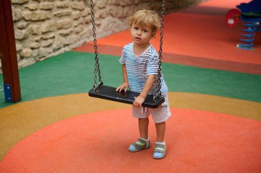 Gündelik giysiler içinde sevimli genç bir çocuk açık havada, yumuşak döşemeli bir parkta salıncağın yanında duruyor. Resim, güvenli ve renkli bir oyun alanındaki eğlence ve çocukluğu resmediyor..