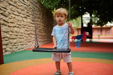 Genç bir çocuk, oyun yapıları ve yeşilliklerle çevrili, yumuşak zemini olan canlı bir oyun parkında salıncağın yanında duruyor. Sahne, dışarıdaki bir keşif ve çocukluk aktivitesini resmediyor..