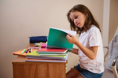 Genç bir kız bir masada oturur, defterleri karıştırır ve ders çalışır. Çeşitli kırtasiye malzemeleriyle çevrelenmiş olarak, öğrenme ve akademik başarı elde etmeye odaklanmış durumda..