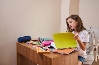 Genç bir kız masasında oturuyor, parlak renkli bir not defterini eleştiriyor. Masa, okul malzemeleri ve malzemeleriyle organize edilerek, odaklanmış ve eğitici bir ortam yaratılıyor..