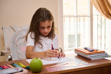 Genç bir kız masasında kitaplar, kalemler ve yeşil bir elmayla çevrili olarak çizim ve yazmaya odaklanıyor. Görüntü, parlak ve rahat bir alanda yaratıcı ve çalışkan bir anı yakalar..